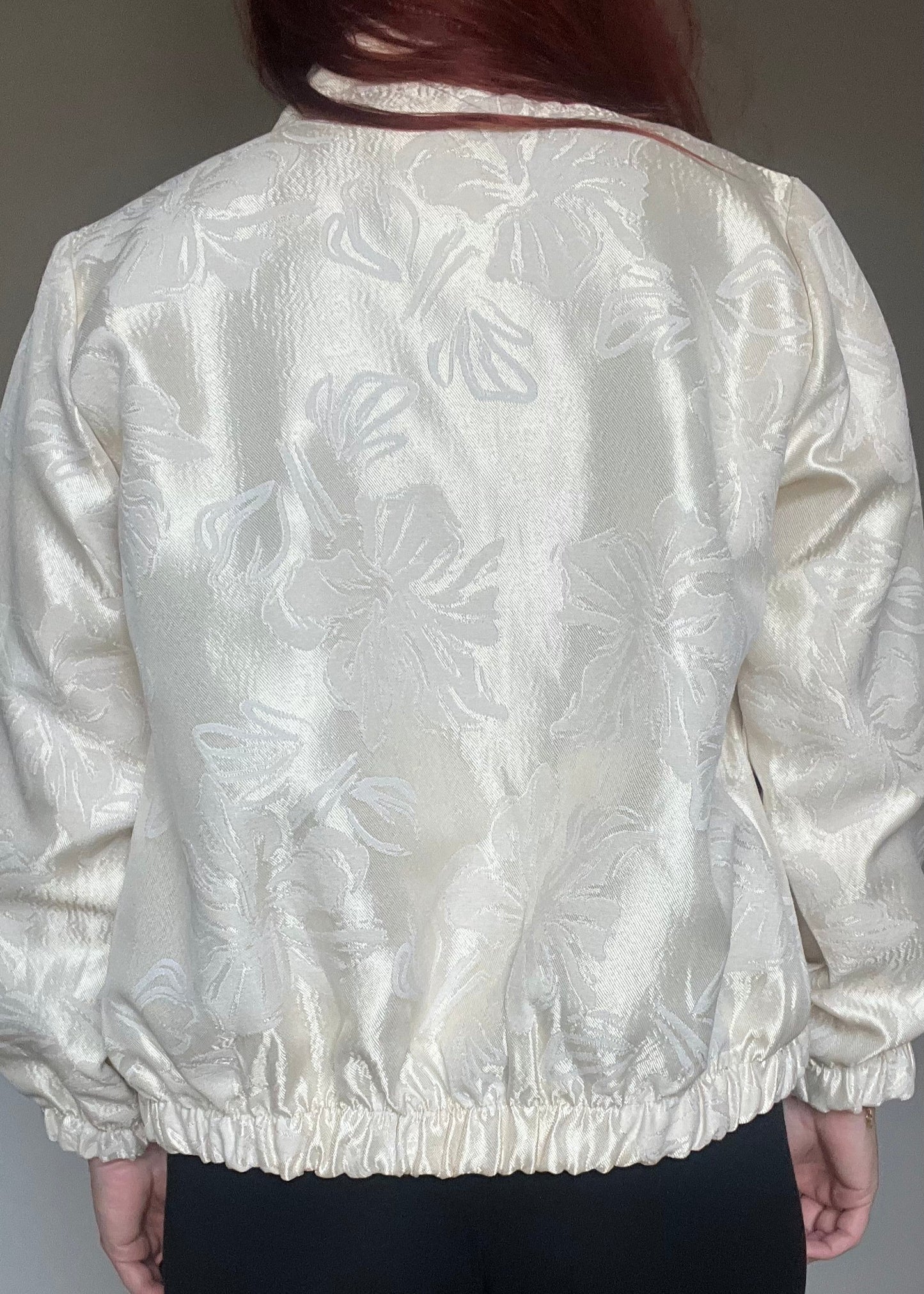 Champagnekleurig semi bomberjasje van glanzende stof met bloemenprint, rits en steekzakken. Vrouwelijk, trendy en one size.