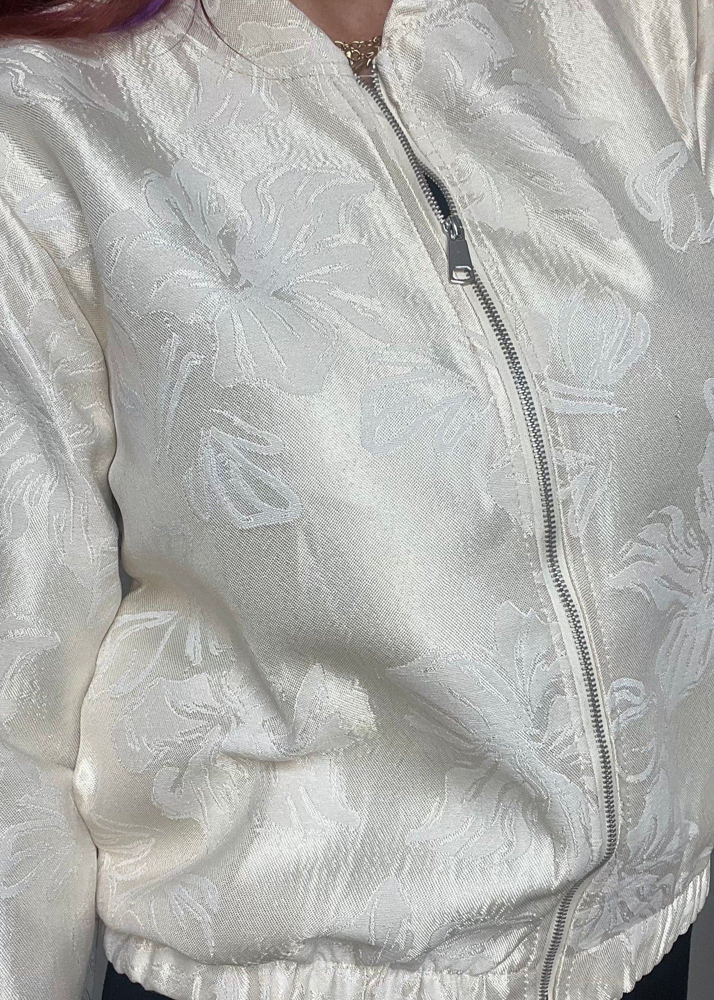 Champagnekleurig semi bomberjasje van glanzende stof met bloemenprint, rits en steekzakken. Vrouwelijk, trendy en one size.